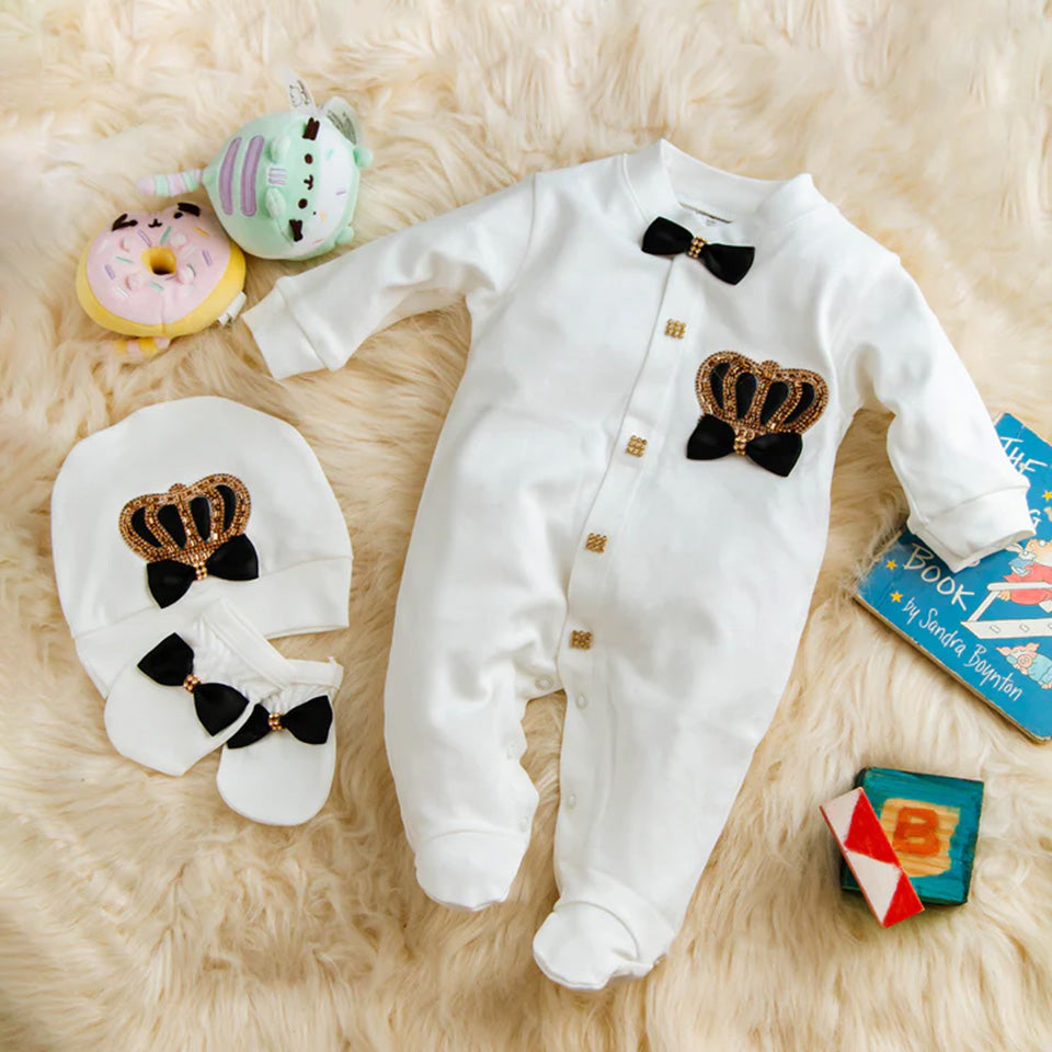 Luxury white baby romper with gold crown and bow tie, matching hat and booties on fluffy rug