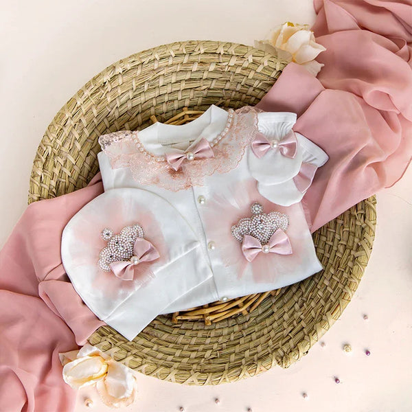 Luxury newborn baby romper with lace, pearls, and pink bows, styled on a woven basket with soft fabric accents.