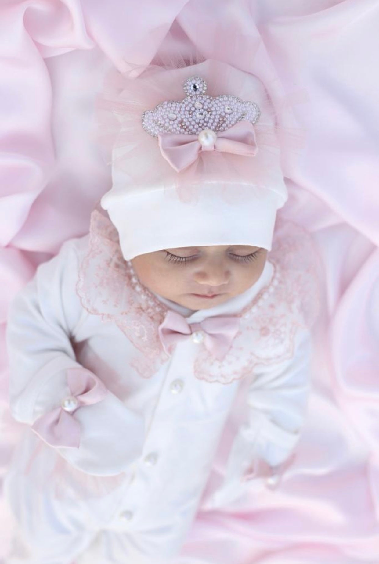 Luxury newborn baby outfit with pink lace, pearl bow, and crown hat on soft pink fabric