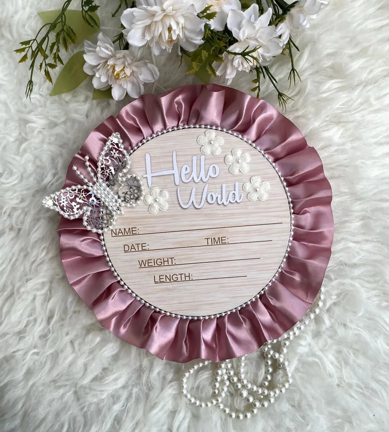 Pink satin 'Hello World' baby announcement plaque with pearl trim, lace butterfly, and floral accents on fluffy white background